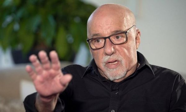 Paulo Coelho sentado com a mão levantada. Ele falou do Talibã