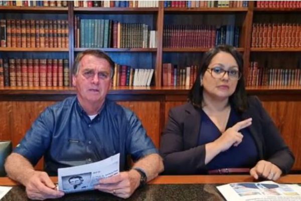 Bolsonaro durante live no YouTube, segurando um jornal com a tradutora de libras ao lado