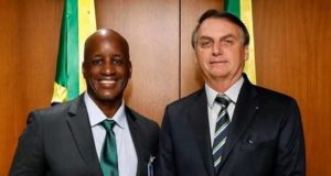 Sérgio Camargo sorrindo, ao lado de Jair Bolsonaro, com bandeira do Brasil atrás. Os dois estão de pé