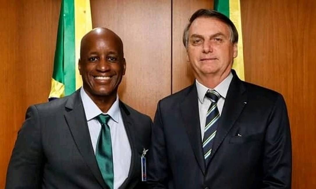 Sérgio Camargo sorrindo, ao lado de Jair Bolsonaro, com bandeira do Brasil atrás. Os dois estão de pé