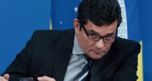 Sergio Moro sentado, segurando celular e olhando por cima dos óculos