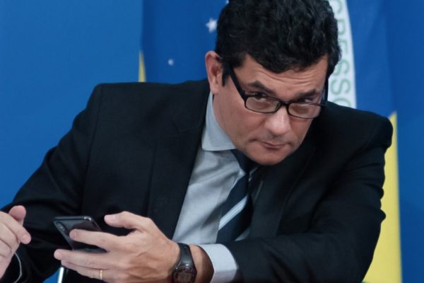 Sergio Moro sentado, segurando celular e olhando por cima dos óculos