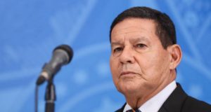 Vice-presidente Hamilton Mourão com olhar sério, usando terno preto e microfone em primeiro plano desfocado.