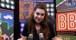 Naiara Azevedo, usando cabelo longo, blusa preta e falando ao microfone em um estúdio.