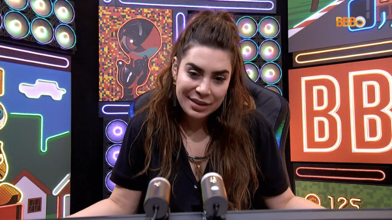 Naiara Azevedo, usando cabelo longo, blusa preta e falando ao microfone em um estúdio.