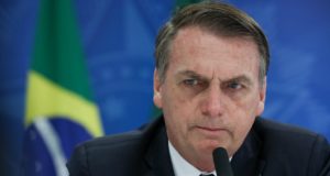 Foto de Bolsonaro falando em microfone. Ao fundo, bandeira do Brasil.