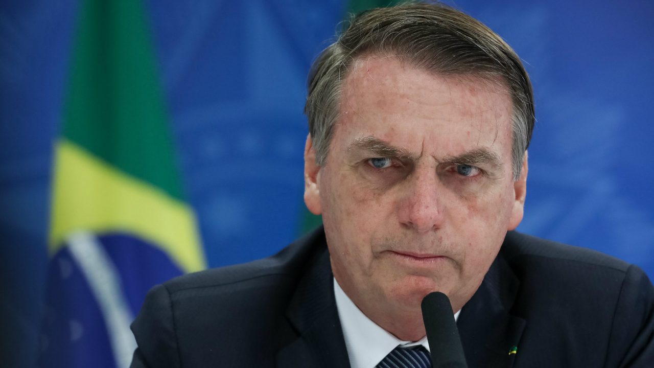 Foto de Bolsonaro falando em microfone. Ao fundo, bandeira do Brasil.