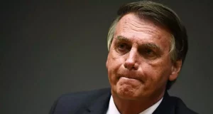 Sentado, Bolsonaro está com a boca fechada e olhar de preocupação, usando um terno preto, camisa social branca e gravata azul. O texto fala sobre o apoio do Centrão ao chefe do executivo federal