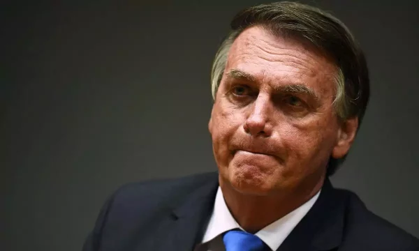 Sentado, Bolsonaro está com a boca fechada e olhar de preocupação, usando um terno preto, camisa social branca e gravata azul. O texto fala sobre o apoio do Centrão ao chefe do executivo federal