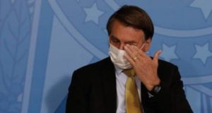 Bolsonaro sentado, de máscaras, e com a mão no olho