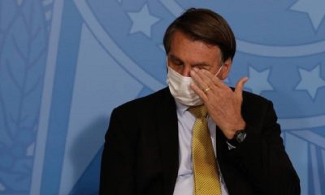 Bolsonaro sentado, de máscaras, e com a mão no olho