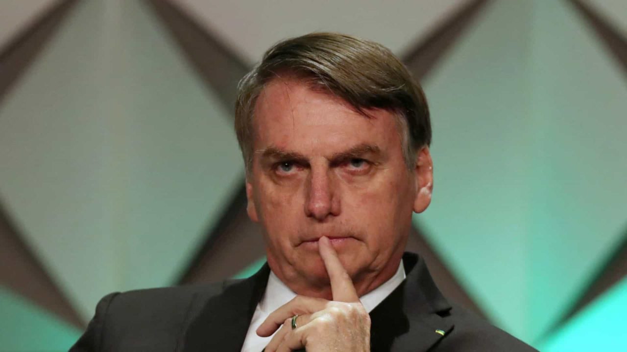 Bolsonaro, sentado e com o dedo na boca, sério