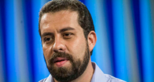 Foto de Boulos usando camisa azul, barba, cabelo curto preto e fundo azul desfocado.