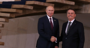 Foto de Putin e Bolsonaro em aperto de mão. Eles estão sorrindo, ambos usam ternos pretos.