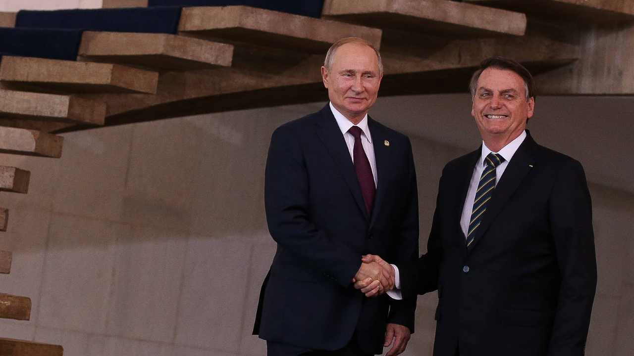 Foto de Putin e Bolsonaro em aperto de mão. Eles estão sorrindo, ambos usam ternos pretos.