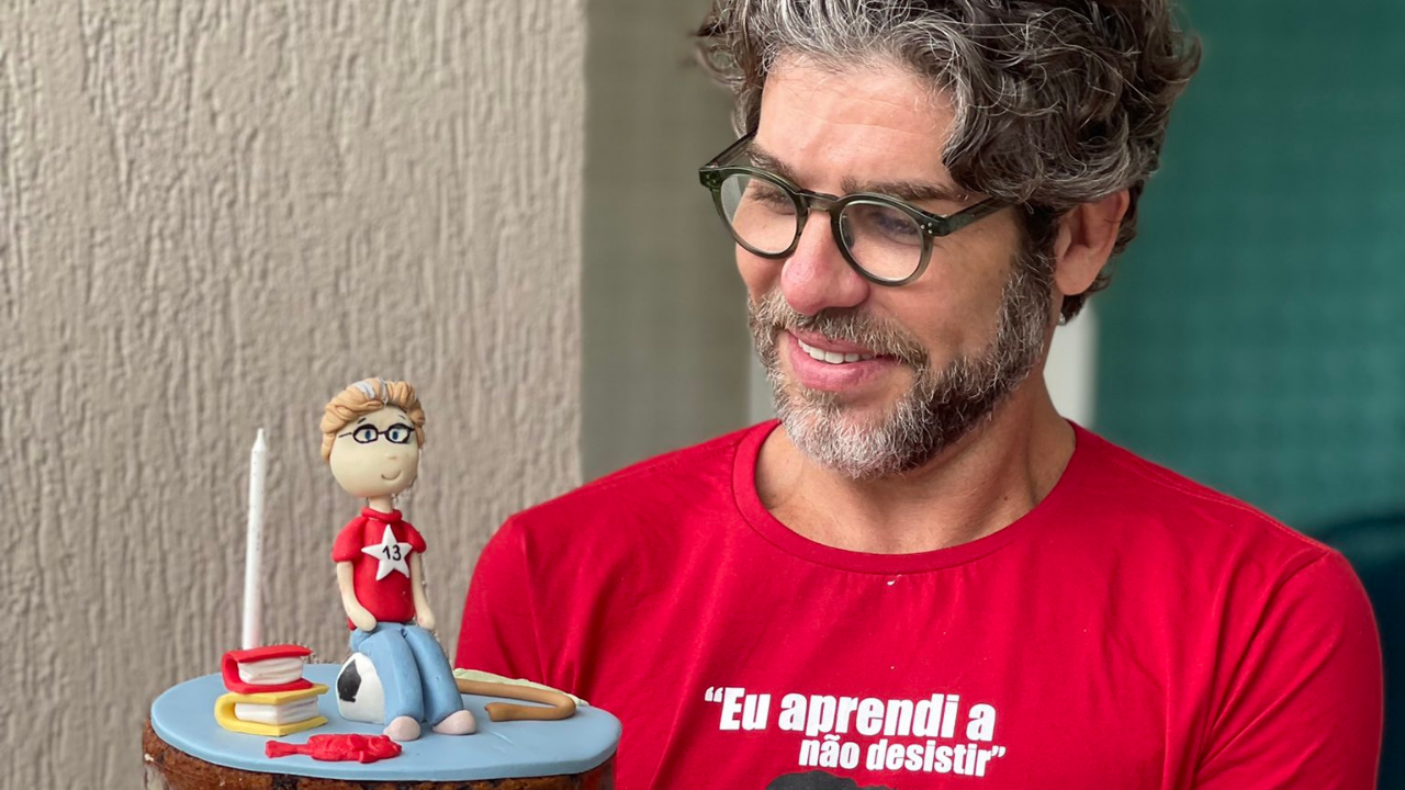 Foto de Juninho Pernambucano com camisa vermelha e bolo com boneco de Lula. Ele tem cabelo e barba grisalhos.
