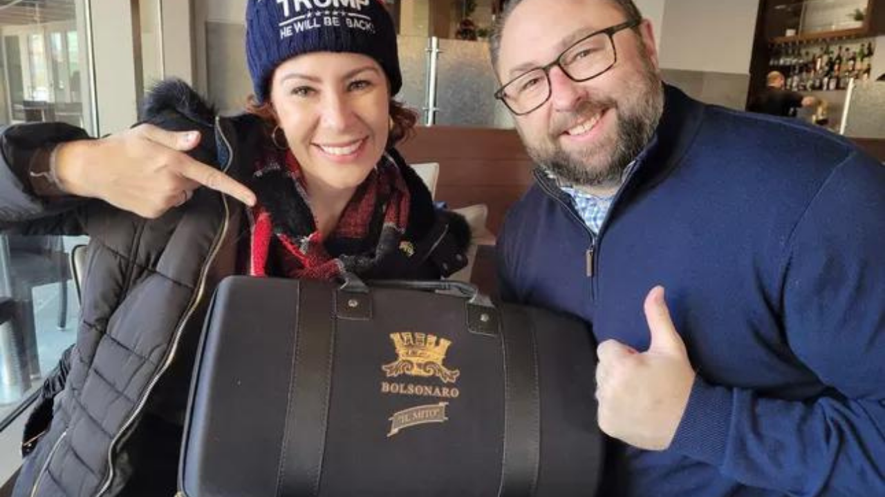 Carla Zambelli usando gorro com o nome Trump e segurando presente para Jason Miller, que está à direita. Ele é branco, tem arba, usa óculos e está com um casaco azul