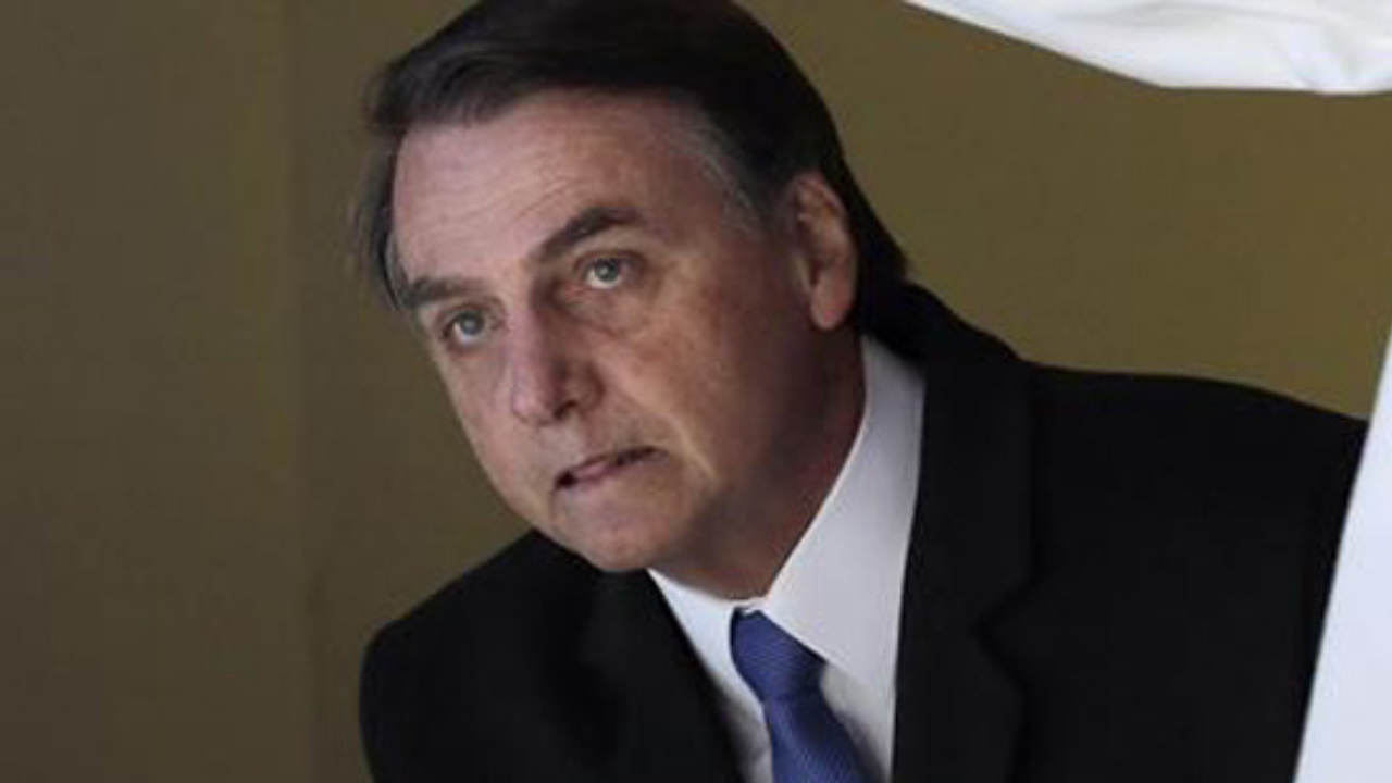 Foto de Bolsonaro com olhar de suspense, esperando algo. Ele usa terno preto com gravata azul.