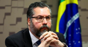 Foto de Ernesto Araújo com olhar pensativo, ele usa óculos e terno preto. Ao fundo, bandeira do Brasil.