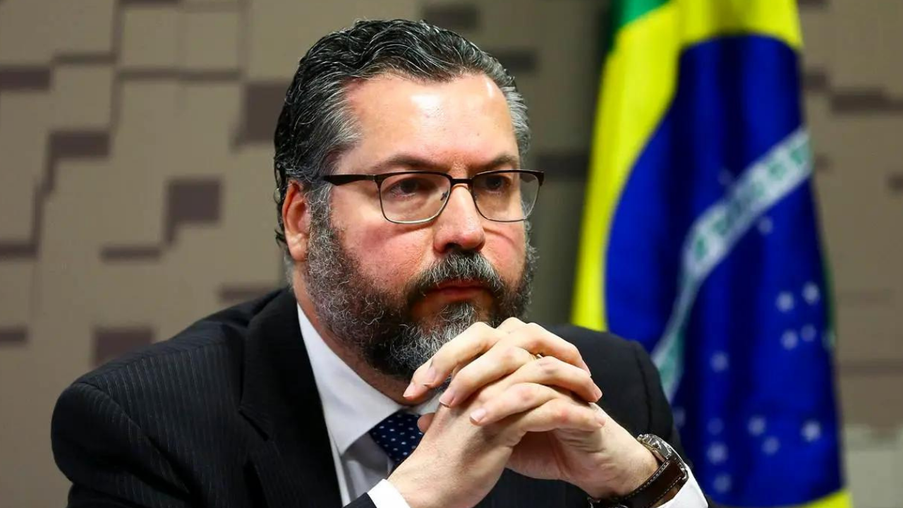 Foto de Ernesto Araújo com olhar pensativo, ele usa óculos e terno preto. Ao fundo, bandeira do Brasil.