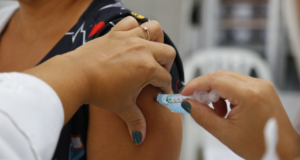 foto de uma pessoa de pele negra recebendo a vacina contra a covid. Na foto, há apenas a imagem do braço direito e uma seringa.