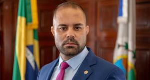 Deputado Filippe Poubel com olhar sério, usando terno e gravata rosa. Ele tem barba e é calvo