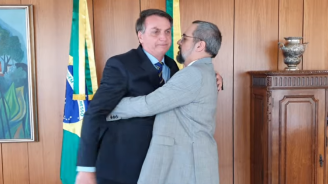 Bolsonaro e Weintraub em um abraço desconcertado no Palácio do Planalto. Ao fundo há bandeiras do país.