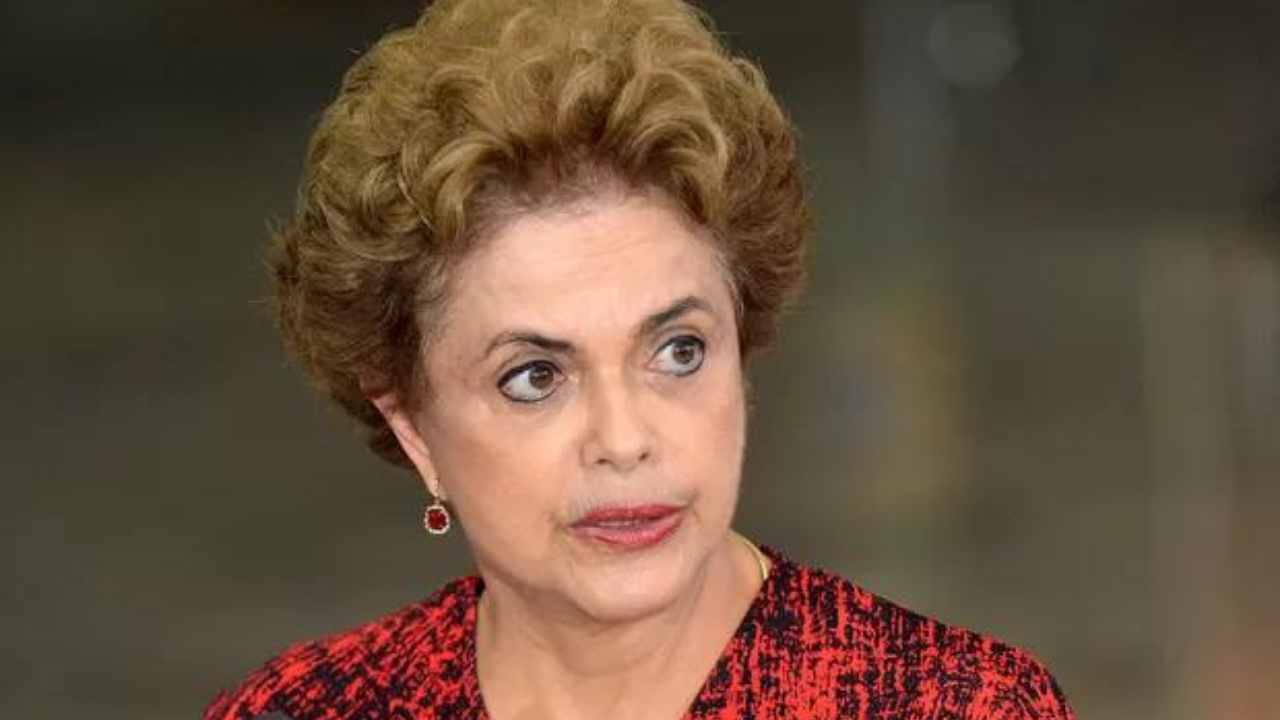 Dilma Rousseff com cabelo castanho claro, brincos vermelhos, batom vermelho e um vestido vermelho com preto.