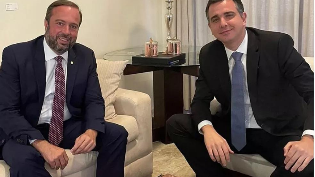 Foto de Alexandre Silveira e Rodrigo Pacheco juntos. eles estão sentados em sofá branco, usando ternos azuis e sapatos pretos. ALexandre usa barba e cabelo grisalho, enquanto Rodrigo tem cabelo preto e não usa barba.