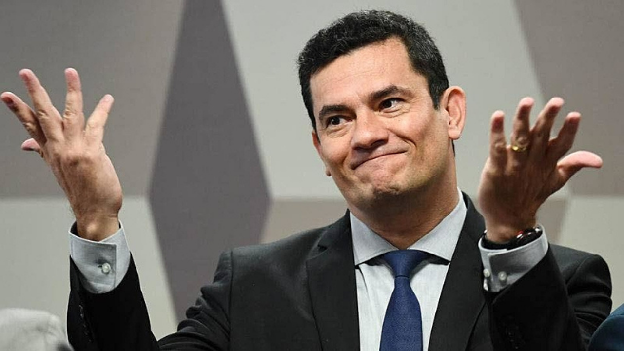 Foto de Moro com as duas mãos ao ar abertas para cima com cara de "não posso fazer nada". Ele usa terno preto e gravata azul, não tem barba e usa cabelo preto.