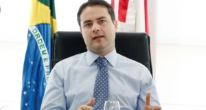 Foto de Renan Filgo com camisa branca, gravata azul com bolinhas e bandeira do Brasil e de Alagoas ao fundo. Ele tem pele branca e cabelo preto.