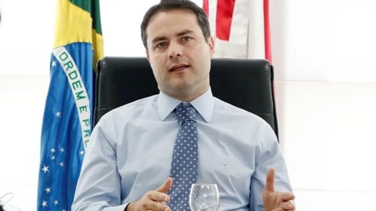 Foto de Renan Filgo com camisa branca, gravata azul com bolinhas e bandeira do Brasil e de Alagoas ao fundo. Ele tem pele branca e cabelo preto.