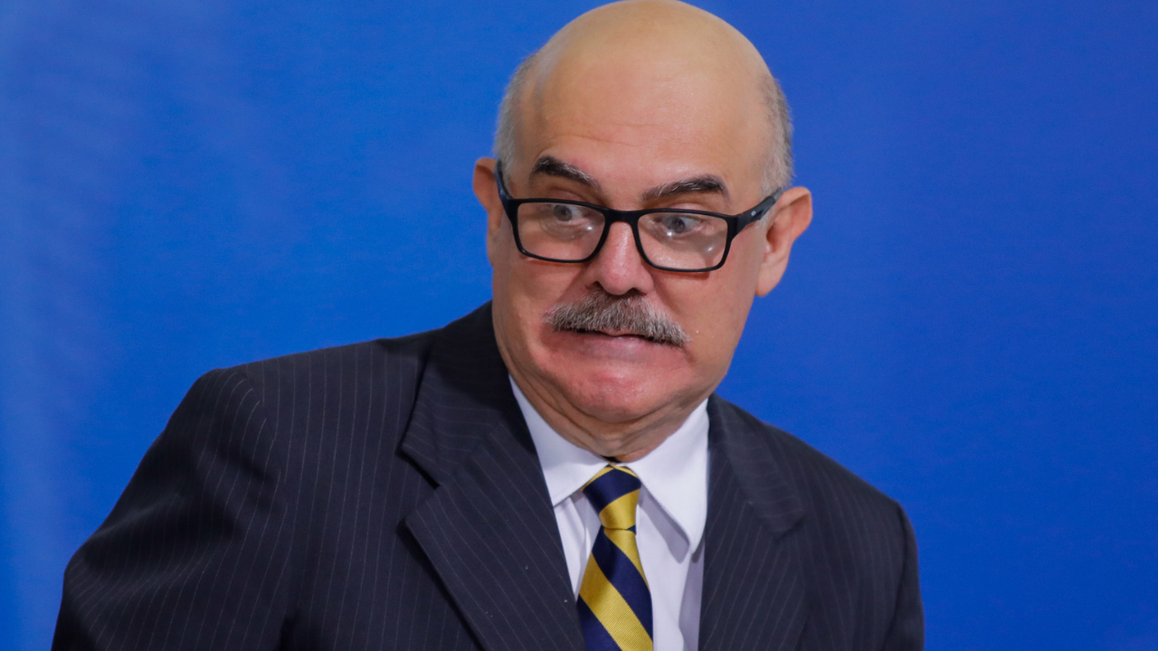 Milton Ribeiro usando terno preto e gravata azul com listras amarelas. Ele usa óculos, bigodo, é calvo e tem cara de susto.