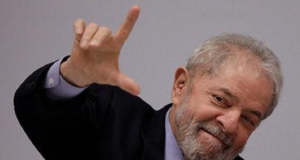 Lula com cabeça inclinada fazendo sinal de L com a mão direita. Ele usa terno preto e tem olhar confiante, fundo branco.