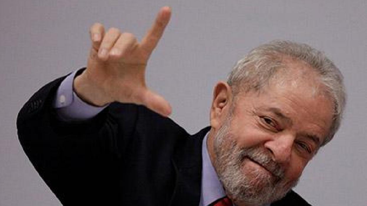 Lula com cabeça inclinada fazendo sinal de L com a mão direita. Ele usa terno preto e tem olhar confiante, fundo branco.