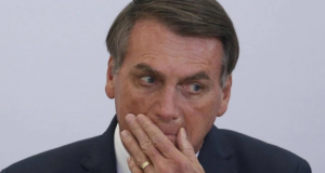 Bolsonaro com olhar preocupado. Ele usa terno preto com a mão esquerda na boca.