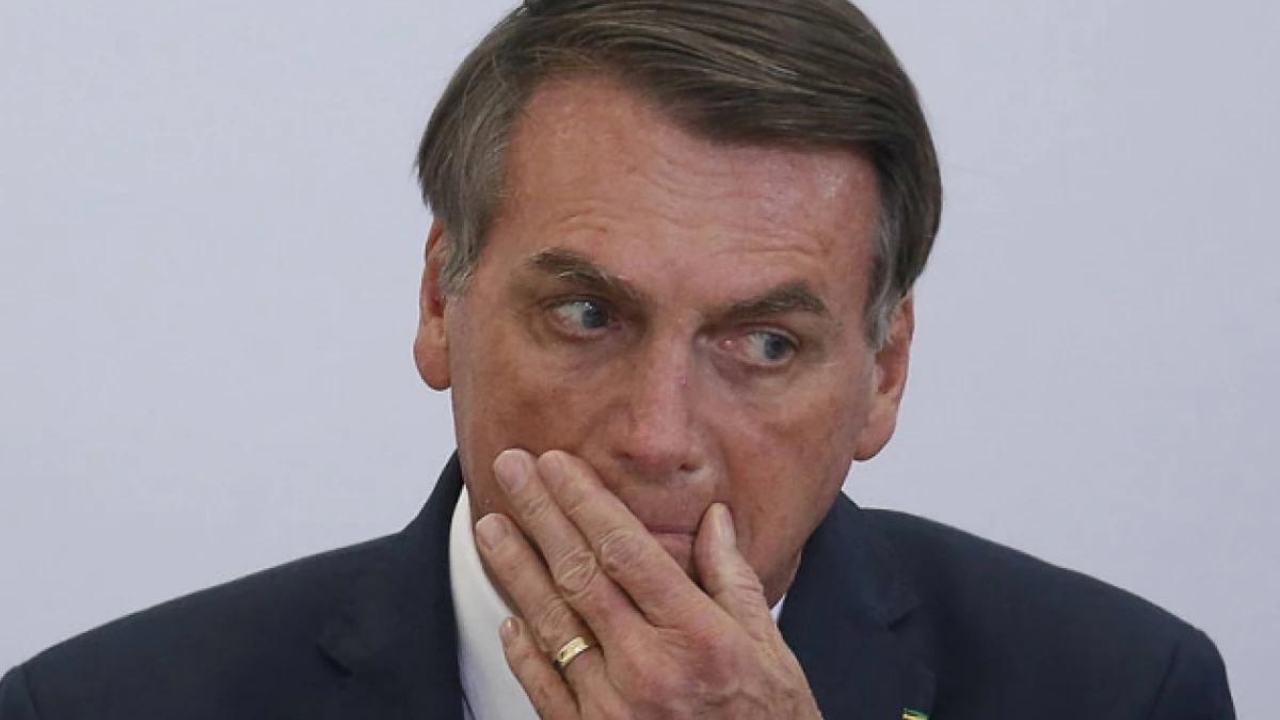 Bolsonaro com olhar preocupado. Ele usa terno preto com a mão esquerda na boca.