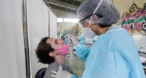 Jovem sentado sendo testado para a Covid-19. Profissional de saúde está de costas para a câmera coletando material.