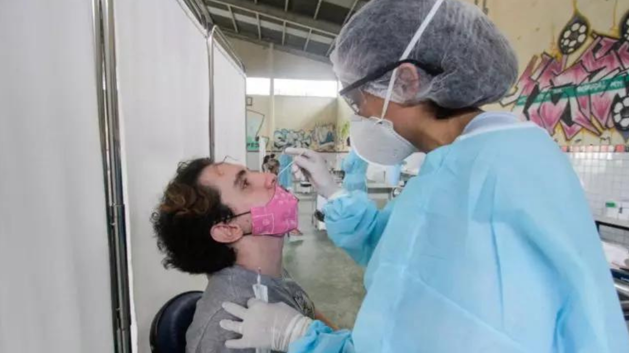 Jovem sentado sendo testado para a Covid-19. Profissional de saúde está de costas para a câmera coletando material.