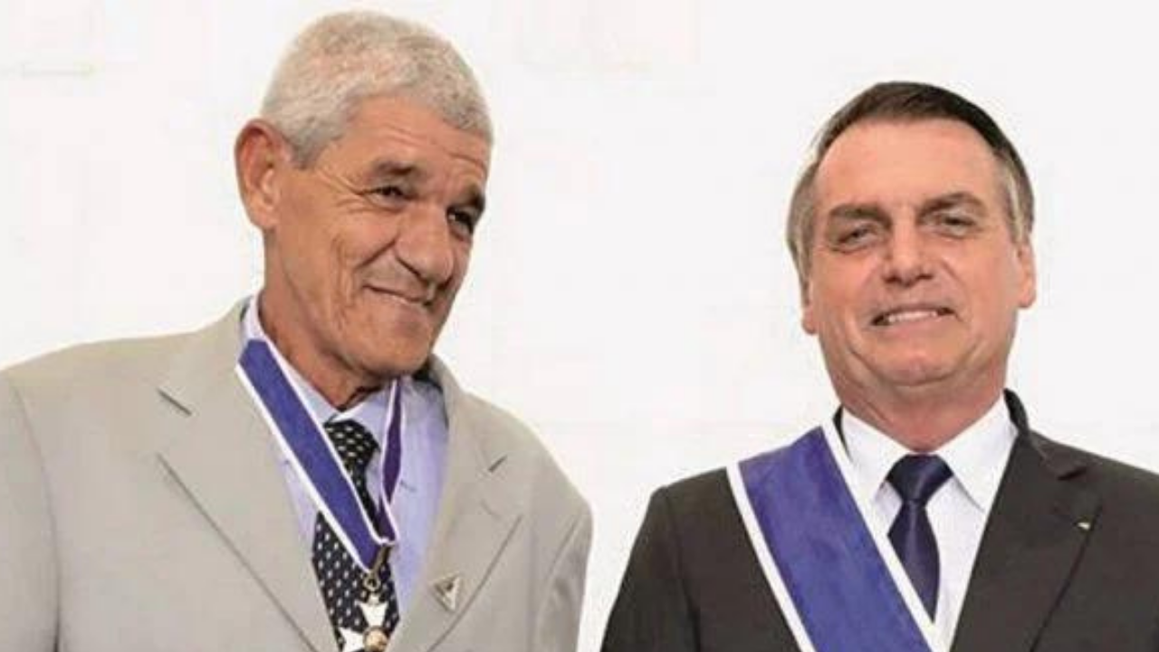Foto de Bolsonaro (à direita) e Ferraz (à esquerda). Bolsonaro usa um terno preto e Ferraz, cujo apelido é Jacaré, usa uma farda do Exército.