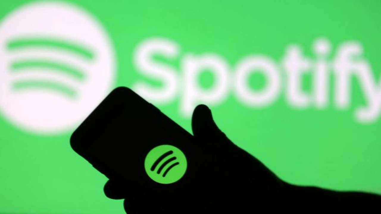 Logo do Spotify ao fundo na cor verde com branco com smartphone na frente mostrando na tela tambéma logo.
