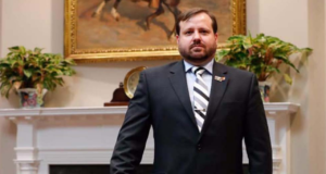 Foto de Célio Faria Junior usando terno e com olhar sério. Ele usa barba e ao fundo há um quadro com um cavalo e um jarrod e flores.