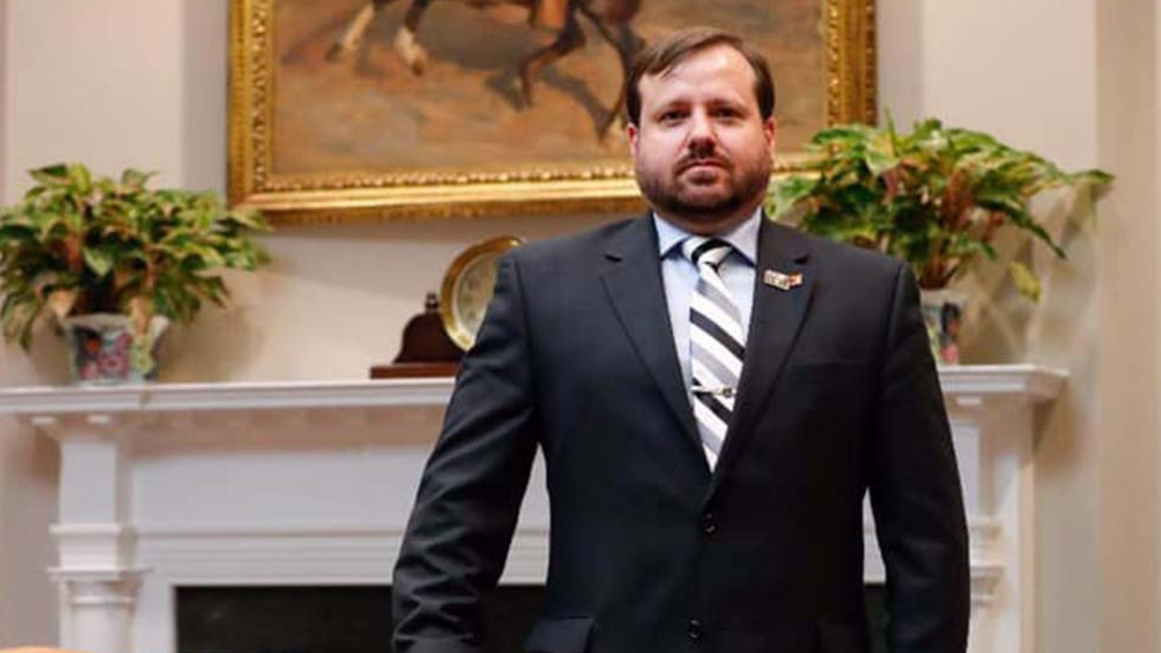 Foto de Célio Faria Junior usando terno e com olhar sério. Ele usa barba e ao fundo há um quadro com um cavalo e um jarrod e flores.