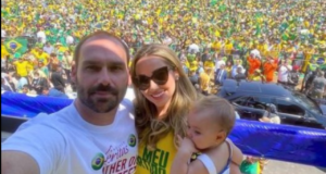 Foto de Heloísa e Eduardo Bolsonaro junto com a filha em um palco. Eles estão em uma manifestação à favor do presidente, ela é loira e usa óculos de sol. Eduardo é calvo e está com uma camisa branca.