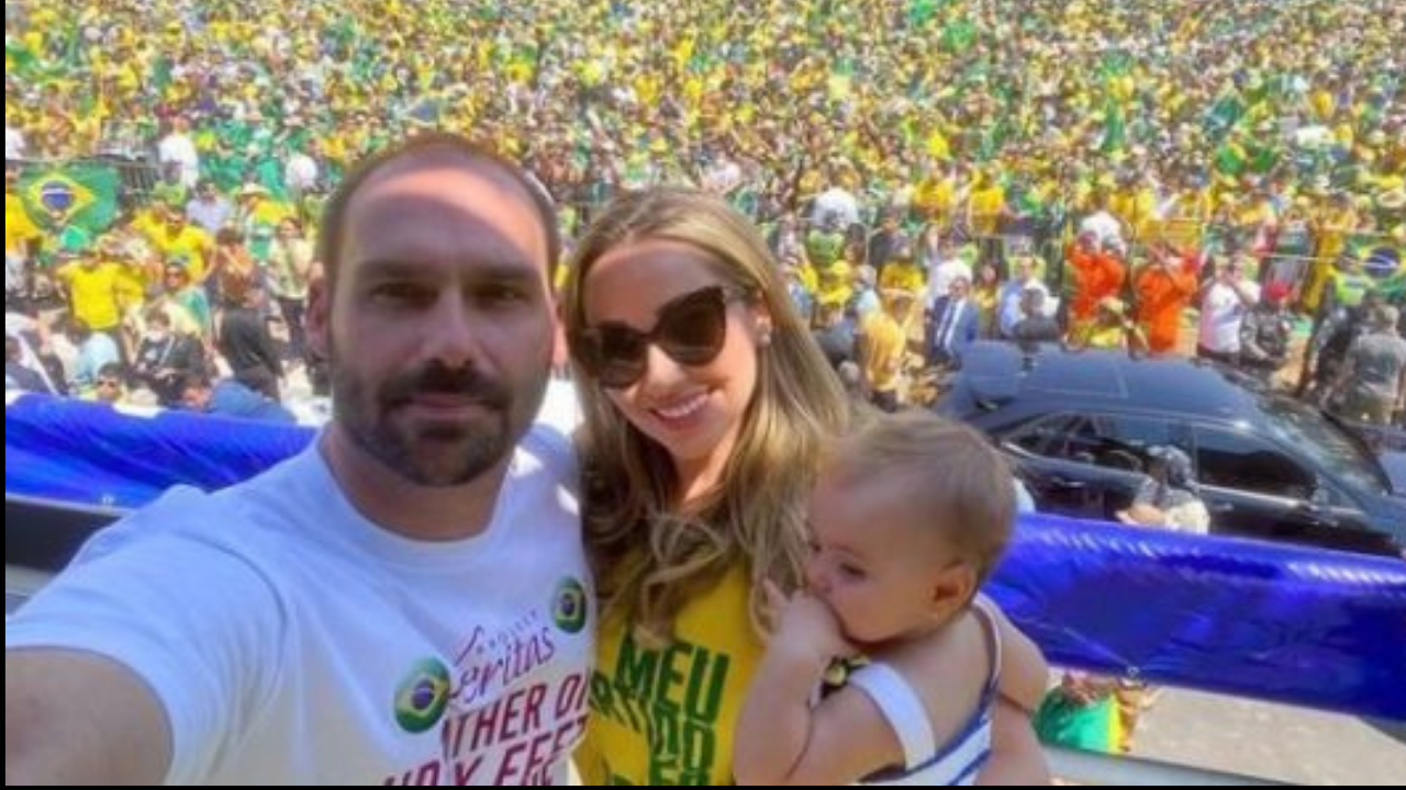 Foto de Heloísa e Eduardo Bolsonaro junto com a filha em um palco. Eles estão em uma manifestação à favor do presidente, ela é loira e usa óculos de sol. Eduardo é calvo e está com uma camisa branca.