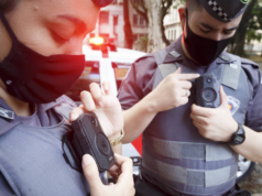Foto de policiais com fardas da PM de Sçao Paulo utilizando câmeras na roupa. Ambos usam máscara e estão com a cabeça abaixada.