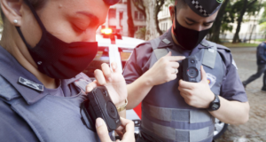 Foto de policiais com fardas da PM de Sçao Paulo utilizando câmeras na roupa. Ambos usam máscara e estão com a cabeça abaixada.