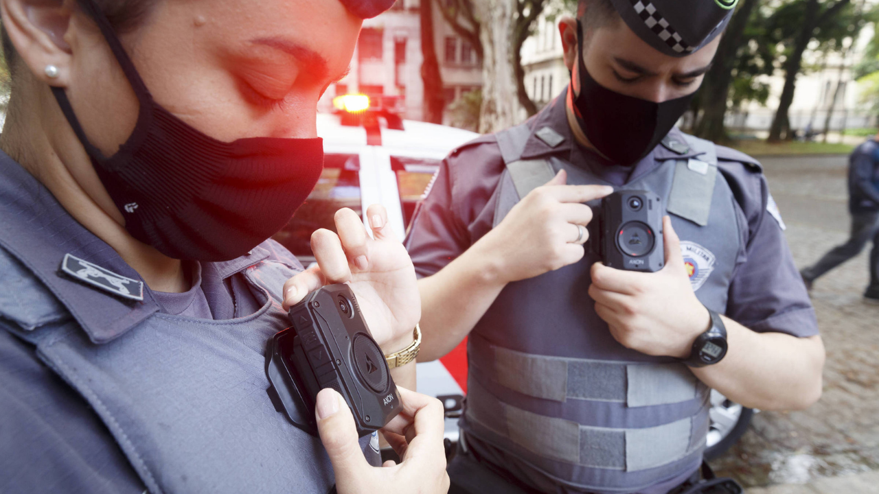 Foto de policiais com fardas da PM de Sçao Paulo utilizando câmeras na roupa. Ambos usam máscara e estão com a cabeça abaixada.