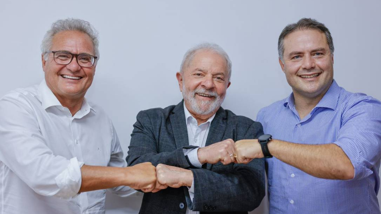 Foto de Renan Calheiros, (à esquerda), com camisa branca, óculos e cabelo branco. Lula ao centro com terno preto, barba e cabelo brancos. Renan Filho à direita, com camisa azul, sem barba e cabelo grisalho.