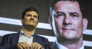 Foto do ex-juiz Sérgio Moro com olhar confiante. Ao fundo uma imagem dele de campanha.